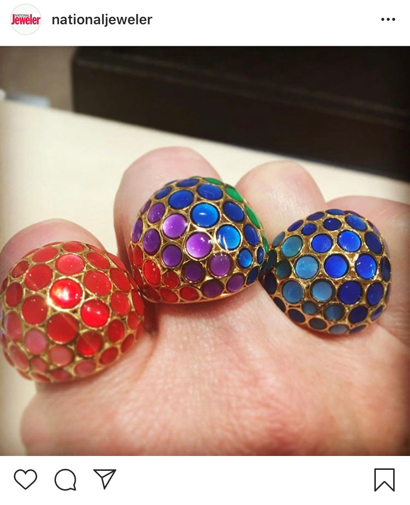 Instagram post from the National Jeweler featuring the Orb Mother of Pearl Gemstone and Enamel 18K Gold Vermeil Sterling Silver Rings. From left to right: tonal red over mother of pearl on gold, rainbow over mother of pearl on gold, and tonal blue over mother of pearl on gold. Picture is of a woman's hand wearing the three rings.