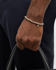 Silver-plated lava stone bead stretch bracelet with oval Jan Leslie script charm pendant worn on wrist with navy trousers and black leather bag