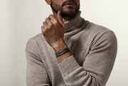 Black wide braided leather bracelet with black steel slide clasp worn on man's wrist with beige turtleneck