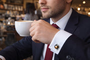 Man in navy suit and red tie holding white coffee cup in café wearing sterling silver spinning globe cufflink with navy blue enamel ocean, multicolor enamel continents, and gold latitude longitude line overlay on white French cuff dress shirt, Jan Leslie.